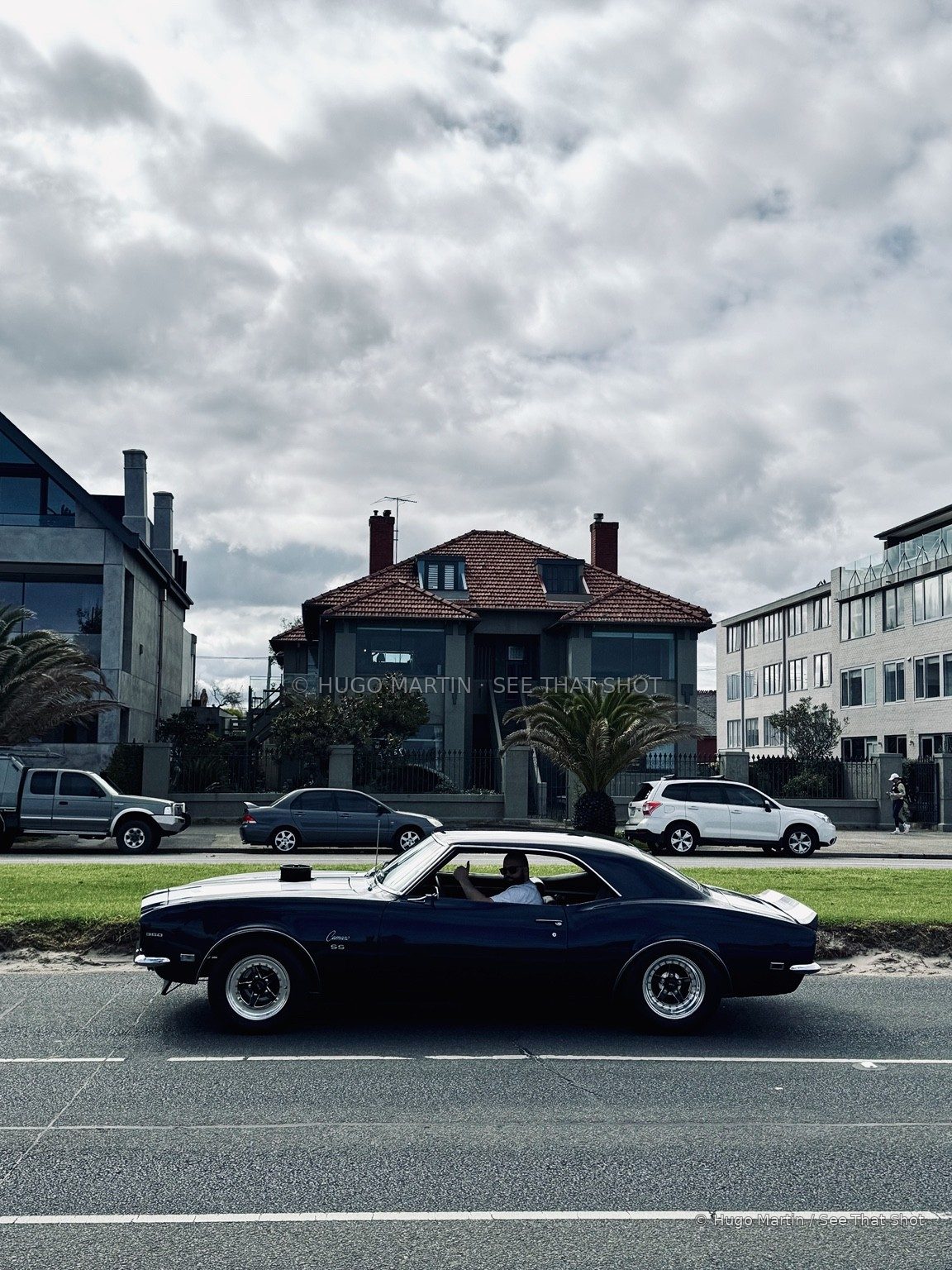 A dark blue vintage Camaro passing beneath a cloudy Melbourne sky